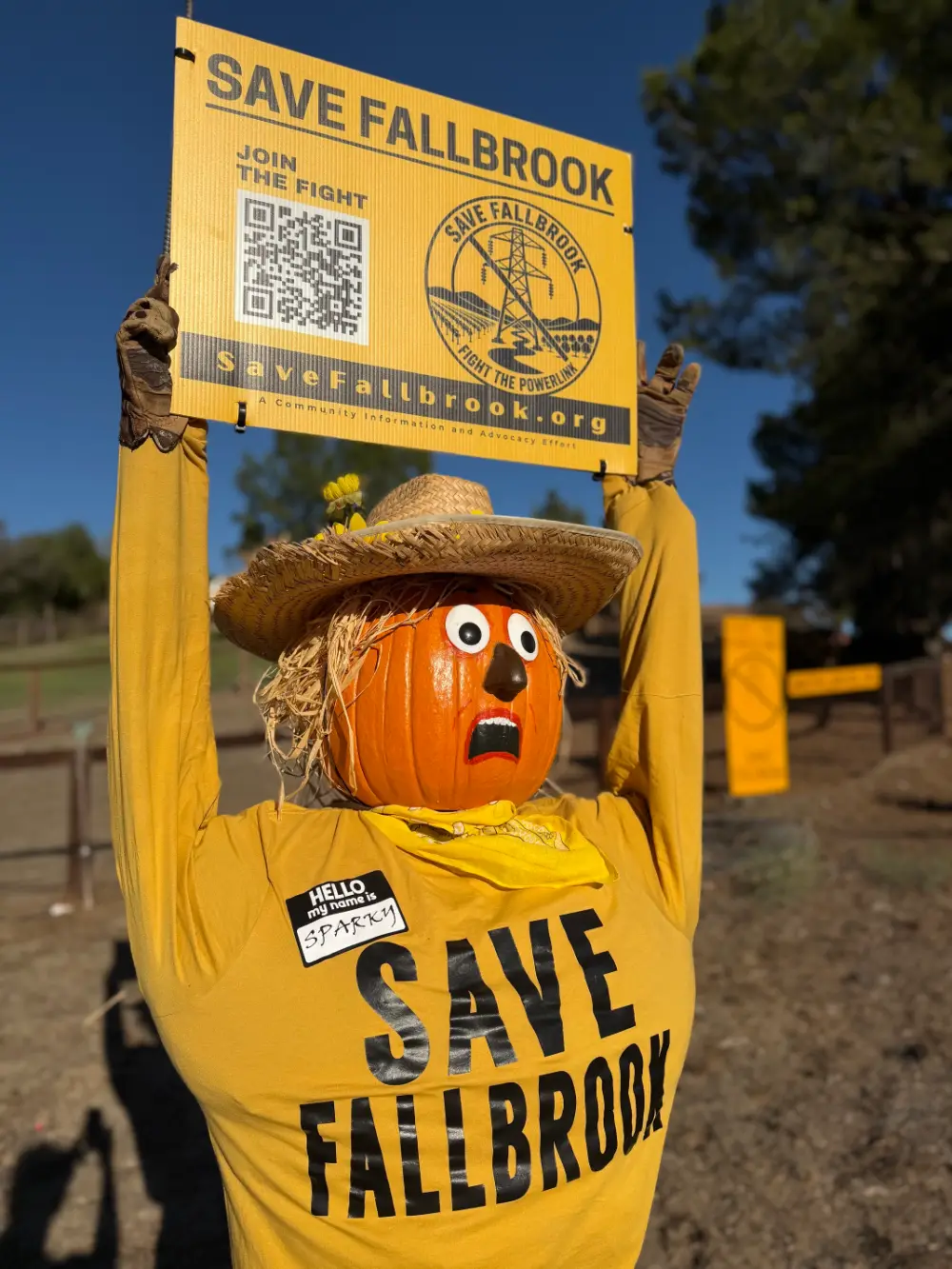 Sparky the Scarecrow, a pumpkin-headed scarecrow in a Save Fallbrook shirt, holding a Save Fallbrook sign over his head