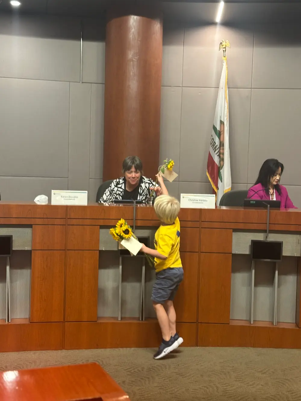 A young Fallbrook boy in a yellow shirt hands a sunflower and a note to Commissioner Karen Douglas at the CPUC meeting